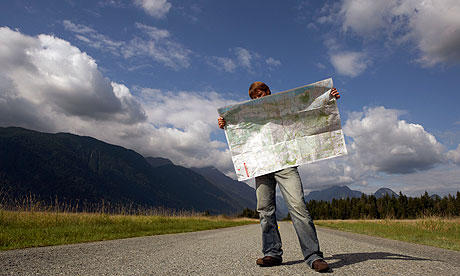 A man standing at the side of a rural road, looking at an unfolded road map.